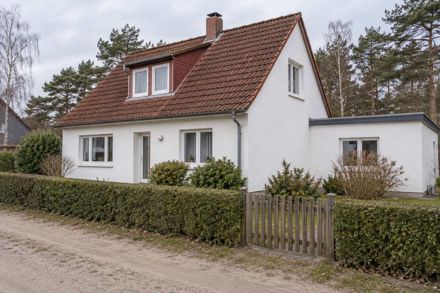 Einfamilienhaus mit weißem Putz, rotem Ziegeldach und Flachdach-Anbau hinter einer Hecke
